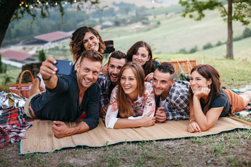 Group of friends taking a selfie during a barbecue in the countryside under a tree - Happy people having fun at a picnic on the hills in summer