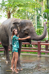 Naklejka premium Smiling Caucasian woman in bright clothes poses next to an elephant, stroking its face with her hand. In the background, a fence and a Park. Vertical