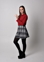 full length portrait of a pretty brunette girl wearing a red shirt and plaid skirt with leggings and boots. Standing pose on a  studio background.