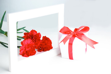 Three red carnation flowers, gift in white packaging with a red bow and frame on a white background. Greeting card for mother's day, international women's day.