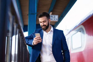 Young bearded businessman in elegant suit waiting for the subway train to get to work and using his...