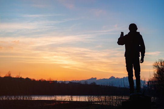 A young man in headphones silhouette looks at the sunset and takes photos on the phone