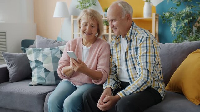 Senior husband and wife are using smart phone in apartment talking swiping screen enjoying modern device. Lifestyle, technology and people concept.