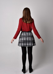 full length portrait of a pretty brunette girl wearing a red shirt and plaid skirt with leggings and boots. Standing pose facing away from the camera on a  studio background.