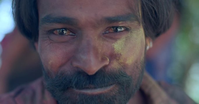 Dark brown Indian man smiling and laughing with big toothy smile intoxicated at Holi festival  colour celebrations- drunk and stoned, with people blurred in background outdoors in Rajasthan, India