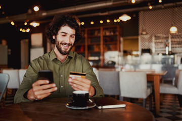 Happy caucasian bearded man holding mobile phone typing card data to make online payment sitting in...