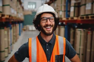 Cheerful male supervisor in warehouse wearing white helmet with orange vest protection suit...