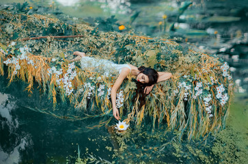 Panele Szklane  River Nymph in white vintage dress lies in boat decorated flowers yellow willow branches, enjoy silence relax. long flowing dark hair. touches hand lily. Backdrop autumn orange nature green water lake
