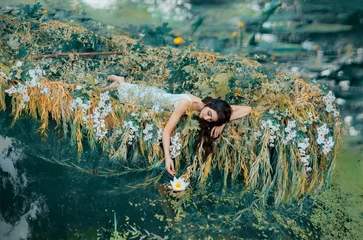Selbstklebende Fototapete Frauen River Nymph in weißem Vintage-Kleid liegt in Boot verzierten Blumen gelbe Weidenzweige, genießen Sie die Stille, entspannen Sie sich. lange fließende dunkle Haare. berührt Handlilie. Hintergrund Herbst orange Natur grüner Wassersee
