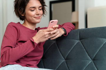 Photo of pleased young woman using smartphone while sitting on sofa