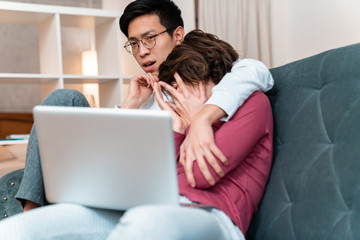 Photo of scared multinational couple hugging and using laptop