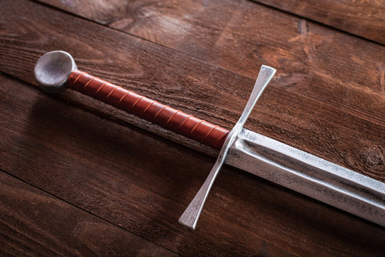 A metal sword on a wooden table with a brown hilt and an unusual guard. Witcher sword made of wood and painted with silver paint. Steel blade.