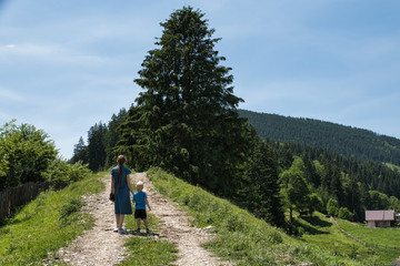 Young mother and little son walking along a dirt trail in nature. Blue sky, hills and forest. Back view.