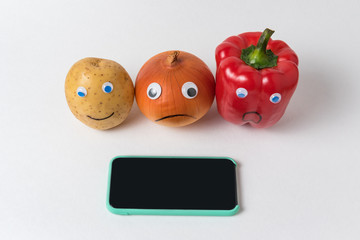Raw potatoes, onions and peppers with funny faces near the mobile phone. App diet concept