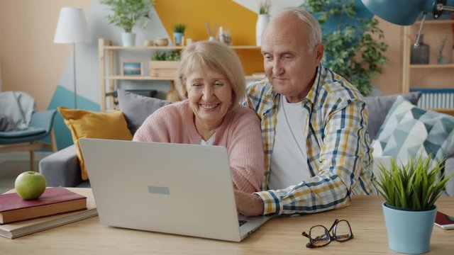 Senior woman and man are using laptop smiling laughing talking looking at screen sitting at table in apartment. People, communication and devices concept.