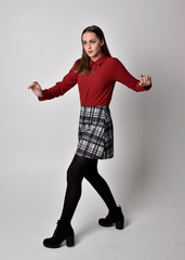 full length portrait of a pretty brunette girl wearing a red shirt and plaid skirt with leggings and boots. Standing pose with hand gesture against a  studio background.