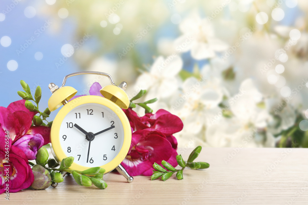 Poster Alarm clock and flowers on wooden table against blurred background, space for text. Spring time