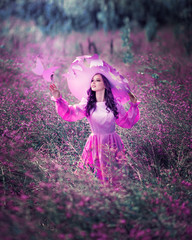 Portrait of amazing cute young woman in beautiful fairy tale image in purple fluffy long dress and with big hat stands in field with purple flowers and stretches her hand to paper purple butterfly