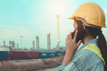 Obraz premium Engineer female or Asian worker with safety helmet hat talking mobile phone while standing looking to factory on Industrial view at oil refinery plant form industry zone. environment, start up concept