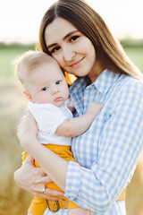 a young happy mother with a small child in her arms, gently leaning against his head, stands in the middle of a field near a collected haystack in the rays of the sunset, summer sun