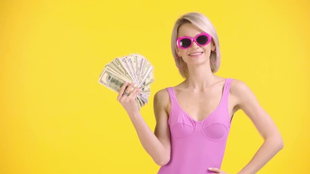 happy girl in swimsuit and sunglasses waving money isolated on yellow