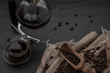 Fototapeta na wymiar Cup of coffee and coffee beans roating with old wooden scoop and coffee beans around on the wooden and dark stone background. Oblique view from the top with copy space for your text.