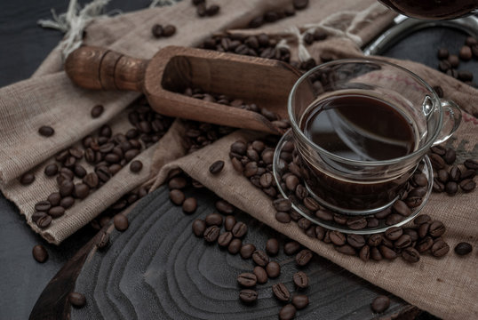 Cup of coffee and coffee beans roating with old wooden scoop and coffee beans around on the wooden and dark stone background. Close-up.
