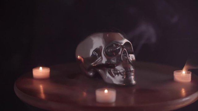 Human chrome skull with candles on a table and smoke, useful for Halloween, or smoking kills message, or displaying a sinister ritual. Downward tilt. 4K