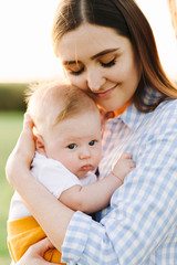 portrait of a young mother in a blue checked dress, holding a small child to her chest, in the rays of the setting summer sun