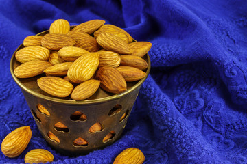 A metal bowl filled with almonds stands on a blue material with a pattern