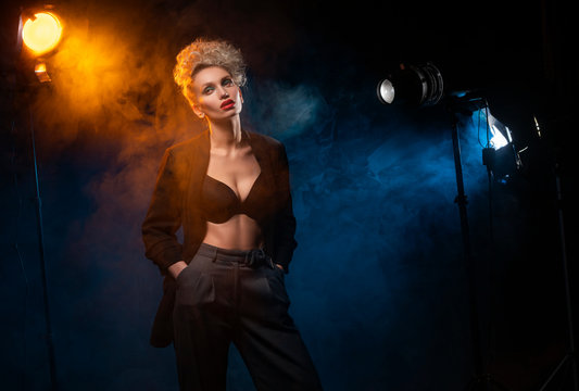 A beautiful blonde girl with an elegant hairstyle and large breasts, wearing a bra, trousers and a blazer, artistically poses in the rays of spotlights in the smoke. Cinematic, art, commercial design.