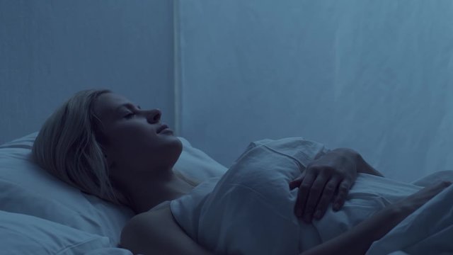 Young woman lying in the bed at night and having insomnia disease. Beautiful blond sleeping girl. Twilight in the bedroom, moonlight from the window. Sleeplessness concept.