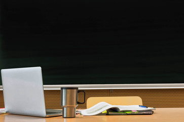 Photo of laptop on professor desk with tumbler and book against black board in classroom
