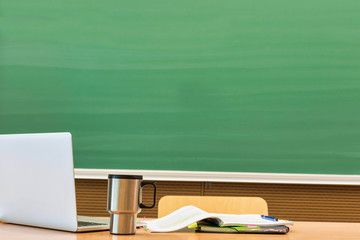 Photo of laptop on professor desk with tumbler and book against black board in classroom