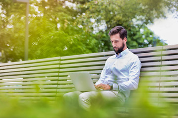 Fototapeta premium Portrait of young attractive businessman sitting on bench while working on his laptop