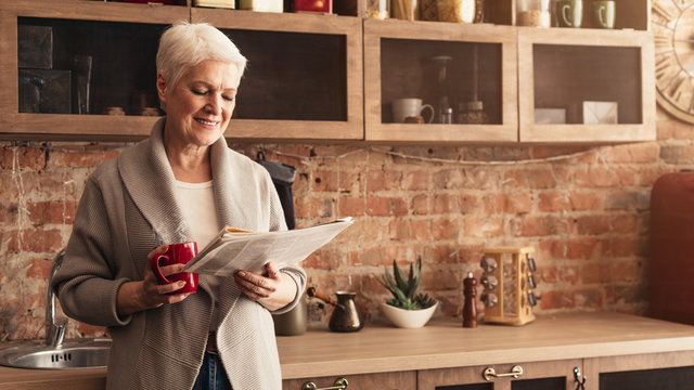 Happy Senior Woman Reading Newspaper And Drinking Hot Tea In Kitchen