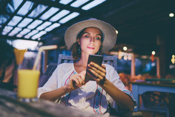 Vacation and technology. Young pretty woman in hat using smartphone sitting at beach cafe bar.