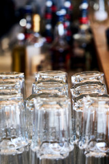 Rows of glasses in a bar waiting to be used. In the background there are bottles of alcoholic beverages.