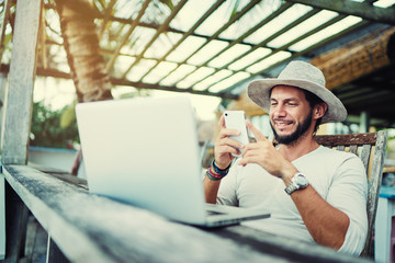 Vacation and technology. Work and travel. Young bearded man in hat using smartphone and laptop computer while sitting at beach cafe bar.