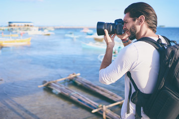 Obraz premium Photography and travel. Young man with rucksack taking photo with his camera on the sea beach near fishing boats.