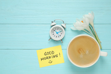 Delicious coffee, flowers and card with GOOD MORNING wish on light blue wooden table, flat lay. Space for text