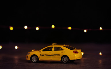 Fototapeta na wymiar Yellow taxi car model on table against festive lights. Bokeh effect