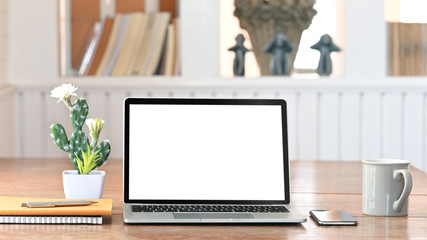 Photo of White blank screen computer laptop, Cactus, Notebook, Pen, Smartphone, Coffee cup flat lay on wooden working table with comfortable living room shelf as background.