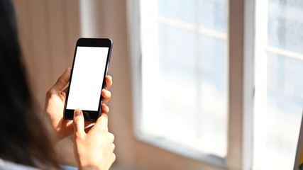 Cropped image back of young woman with long straight hair holding a cropped black smartphone with white blank screen and pressing at the smartphone button with blurred windows as background.