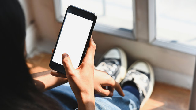 Cropped image of artist woman legs with jeans sitting on wooden floor while holding white blank screen smartphone in hands at the art studio.