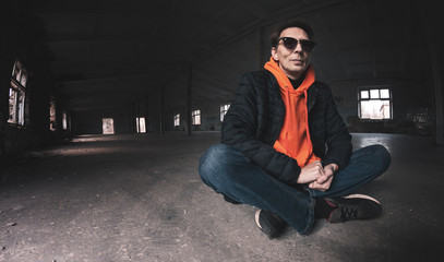 a man in sunglasses sits in a pose with crossed legs on a concrete floor, against the background of an abandoned building with broken windows