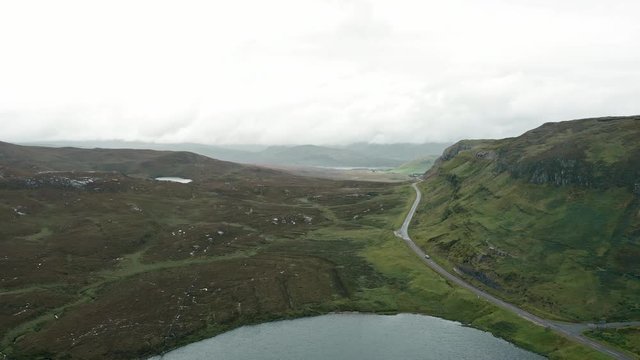 Aerial shot of the rocky ridge of Knockan Crag in the Scottish Highlands. A road curves around the ridge. Peat bogs, grass, marshland and a small lake are in the foreground.