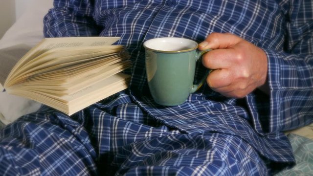 Cinemagraph – Video of steam rising from a hot drink, combined with a still image of a man sitting on a bed, wearing blue patterned pyjamas / pajamas, holding a mug while reading a book. 