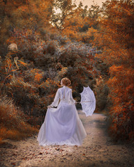 Backside view amazing cute young woman in beautiful fairy-tale image with fire red hair in white long dress holds white lace umbrella and walks along path in bright yellow autumn pictorial forest