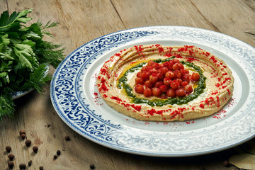 Hummus with red spices, chickpeas and herbs on a ceramic plate on a wooden background. Arabic or Israeli cuisine. Close up, horizontal. Snack